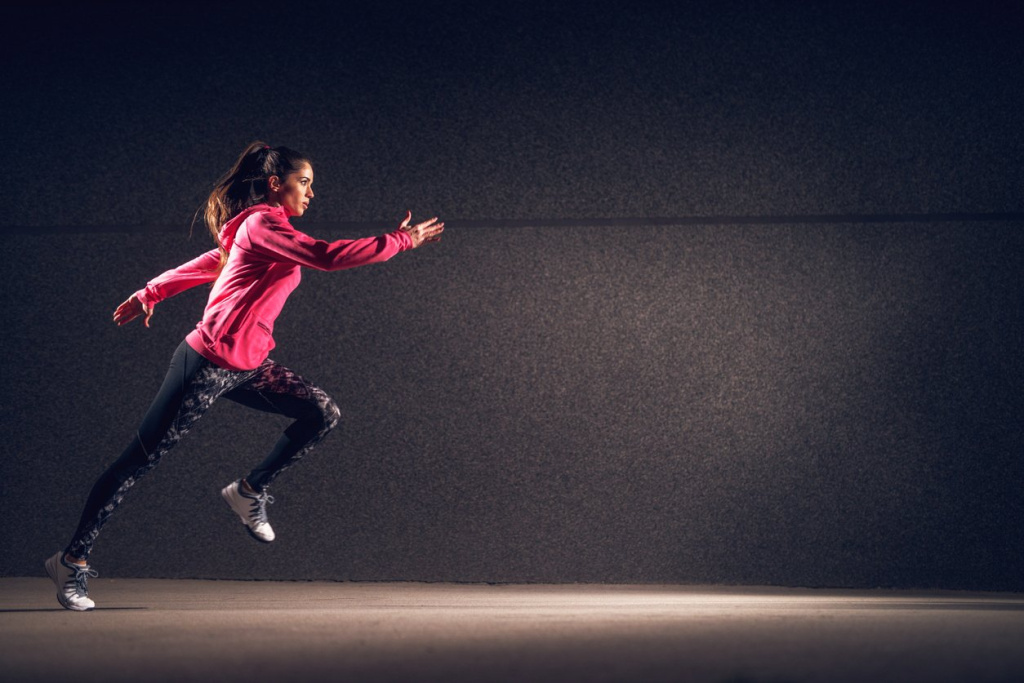 Side motion view of young sexy attractive healthy fitness sporty active slim girl with sportswear running in front of the grey wall at night on the street.